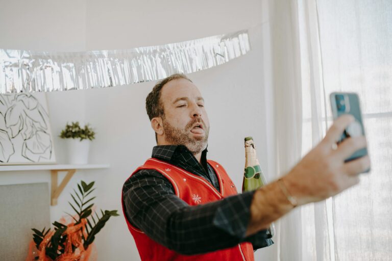 A man in a holiday sweater takes a selfie while holding a beer, capturing a festive indoor scene.